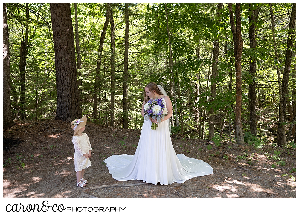 a beautiful Maine bride, standing with her dress flowing around her, smiling at a tiny flower girl wearing a white lace dress, and a straw hat