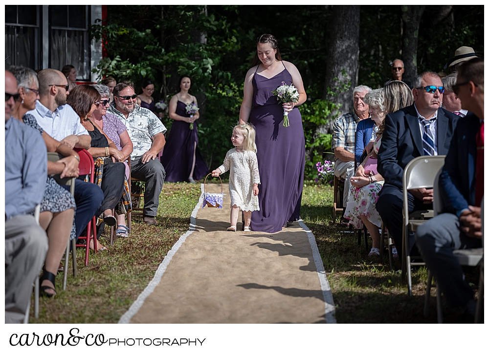 a bridesmaid in purple, walks down a burlap runner, with a tiny flower girl in a white lace dress