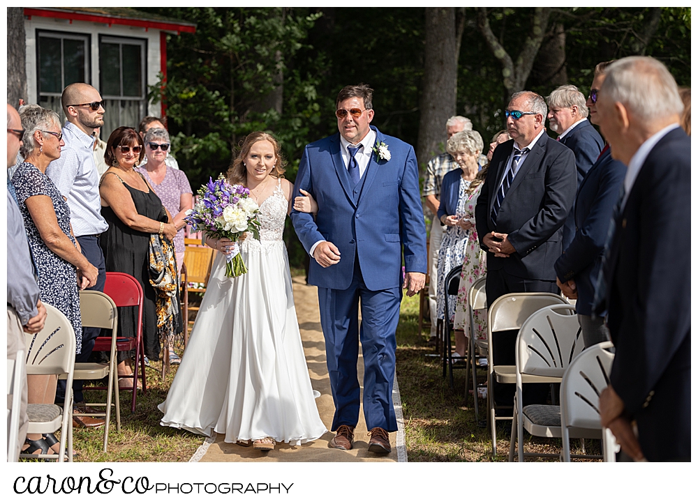 a bride in white, and her dad in blue, walk down the aisle, at a Highland Lake Maine wedding ceremony