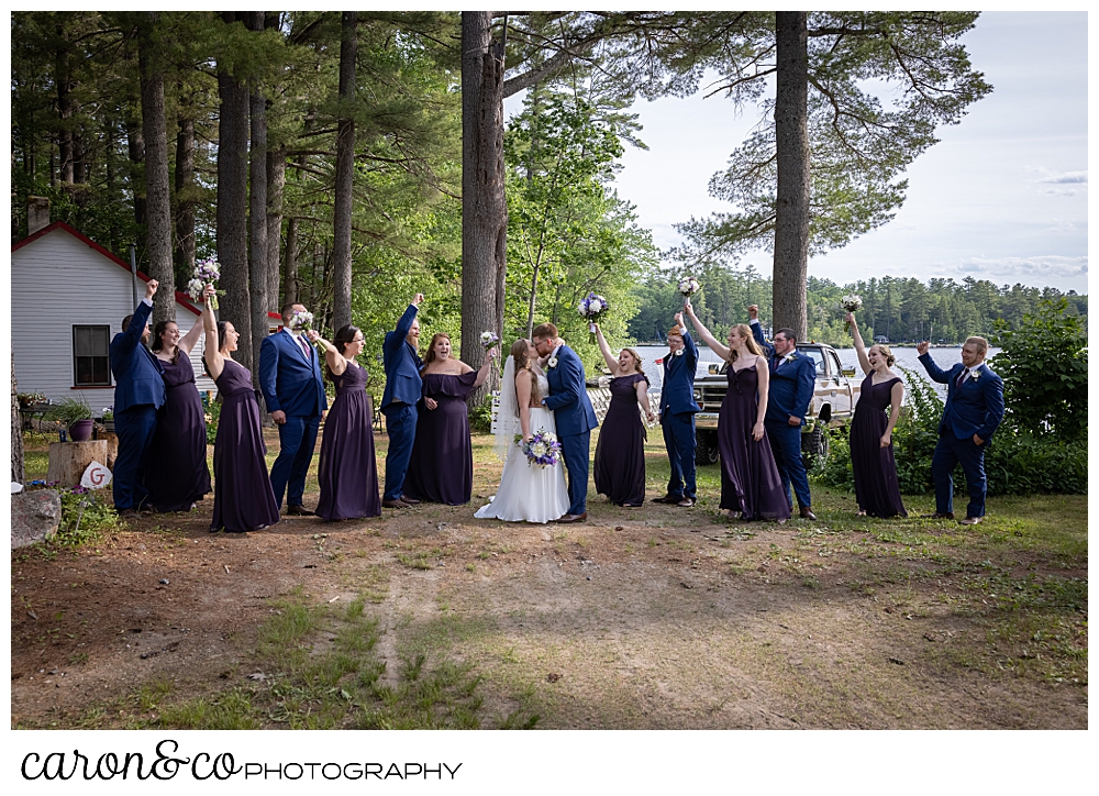 a bride and groom pose with their wedding party, the bridesmaids are dressed in purple, the men in blue, they're loosely grouped together at a highland lake maine wedding ceremony