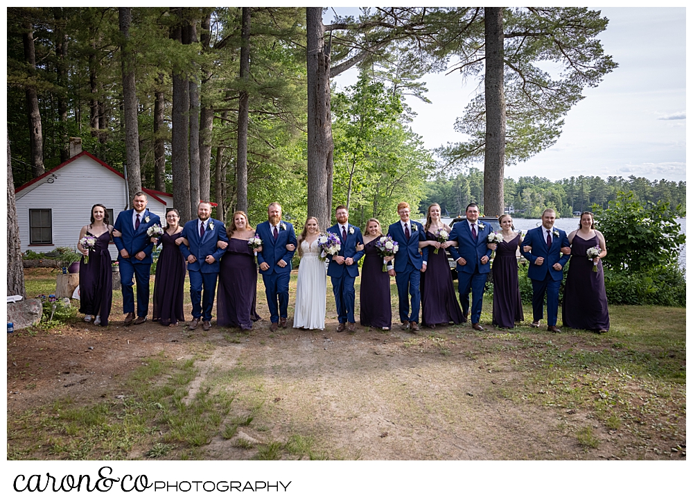 a bridal party with linked arms, walking towards the camera, women are in dark purple, men in blue, at a highland lake maine wedding