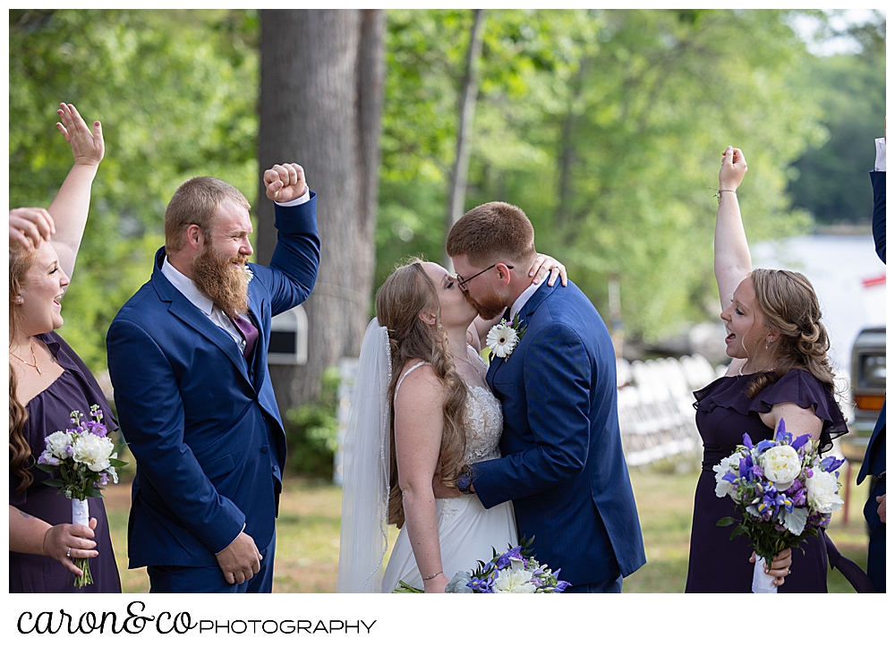 a bride and groom kiss as their wedding party looks on and cheers, at a highland lake maine wedding