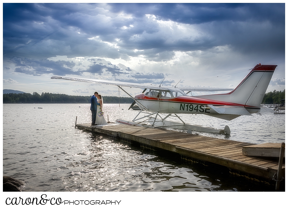 a bride and groom standing on the end of a dock, kissing, with a sea plane in the background, at their highland lake maine wedding