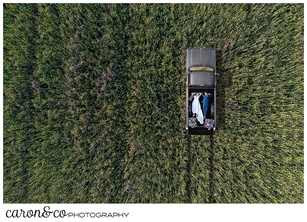 a maine drone wedding photo of a bride and groom lying in the truck bed of an antique truck, in a field of grass and flowers