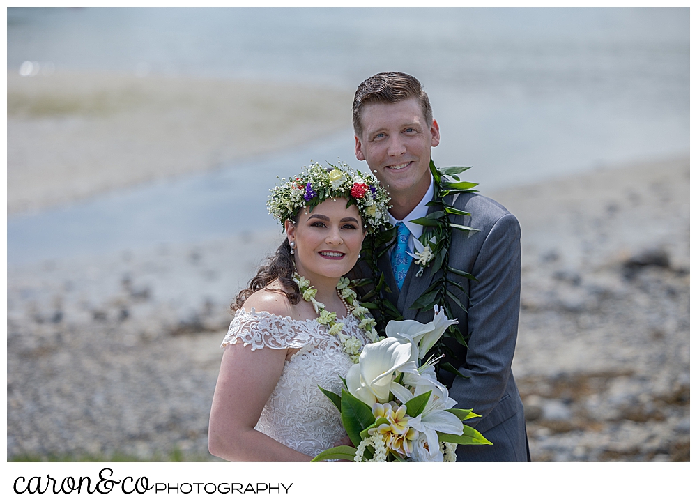 a beautiful portrait of a bride and groom, the bride is wearing a white dress, a flowered headpiece, and a white lei; the groom is wearing a gray suit, a blue tie, and a green lei