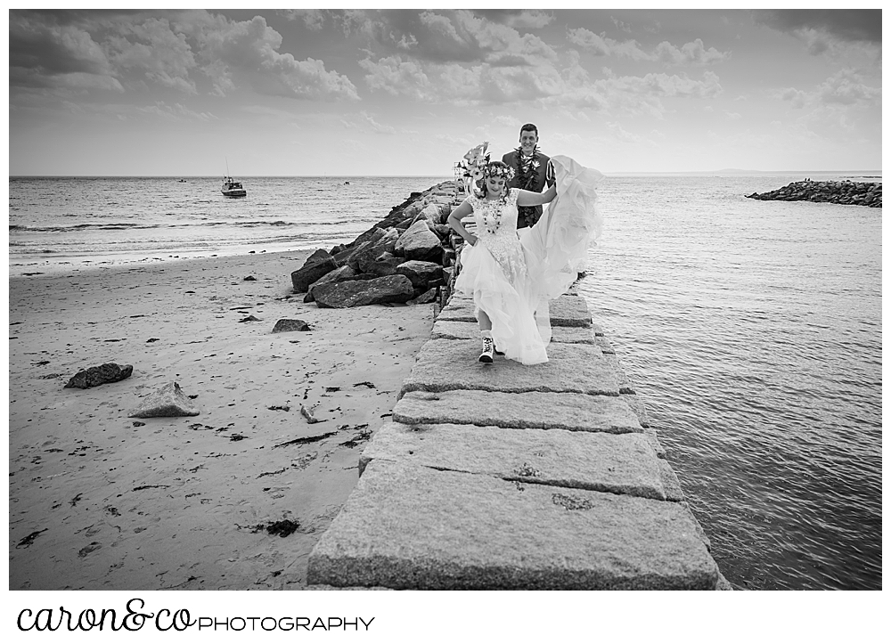 black and white photo of a bride and groom walking on the Kennebunkport Breakwater, the bride is holding her dress