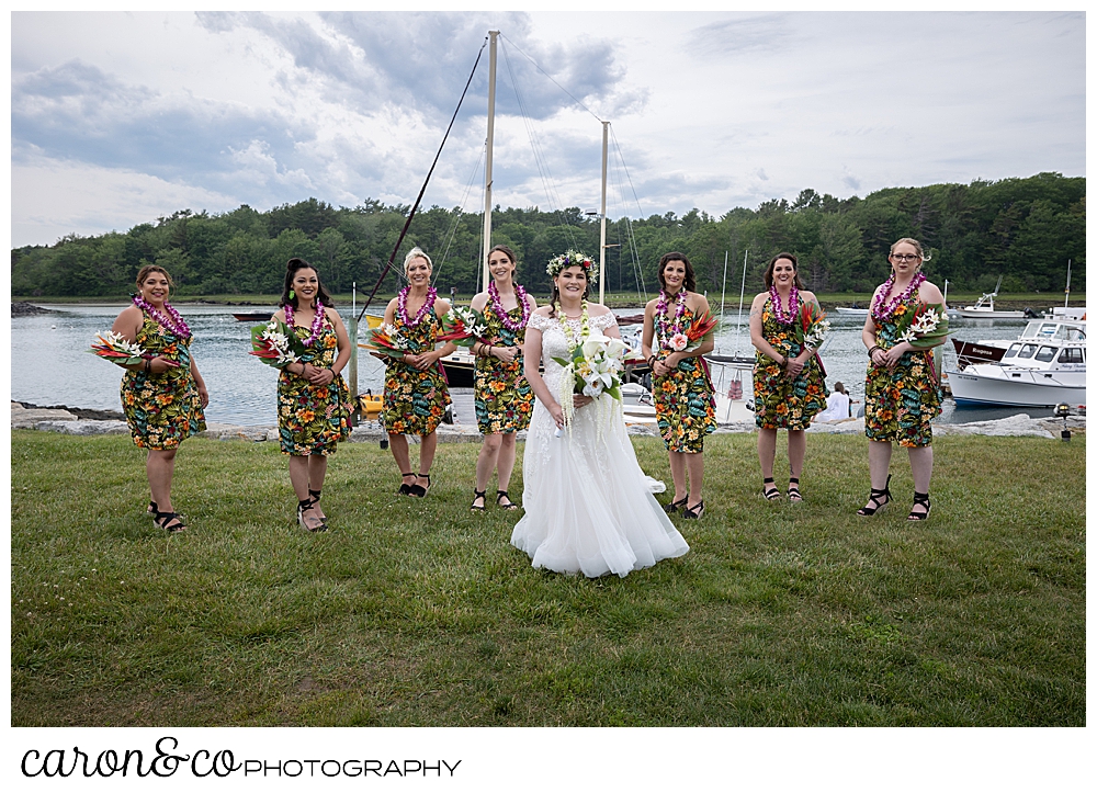 a bride wearing white, is surrounded by 7 bridesmaids wearing short, hawaiian dresses, on the river's edge at a Nonantum Resort tented wedding