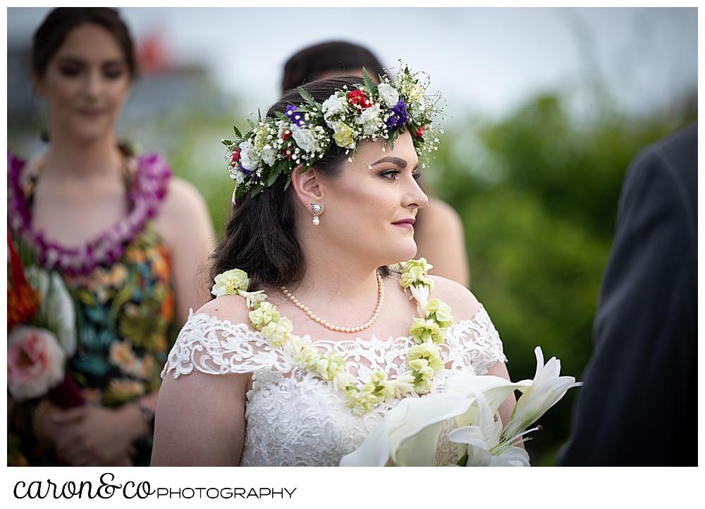 a bride wearing a white dress, a flowered headpiece, and a white lei, looks towards her groom during their outdoor wedding ceremony at the Nonantum Resort Kennebunkport, Maine