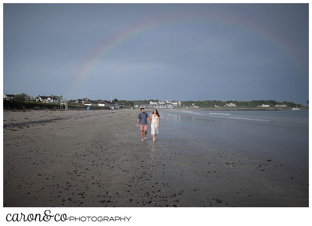 a man and woman walk on Gooch's Beach in Kennebunk, Maine, with a rainbow overhead