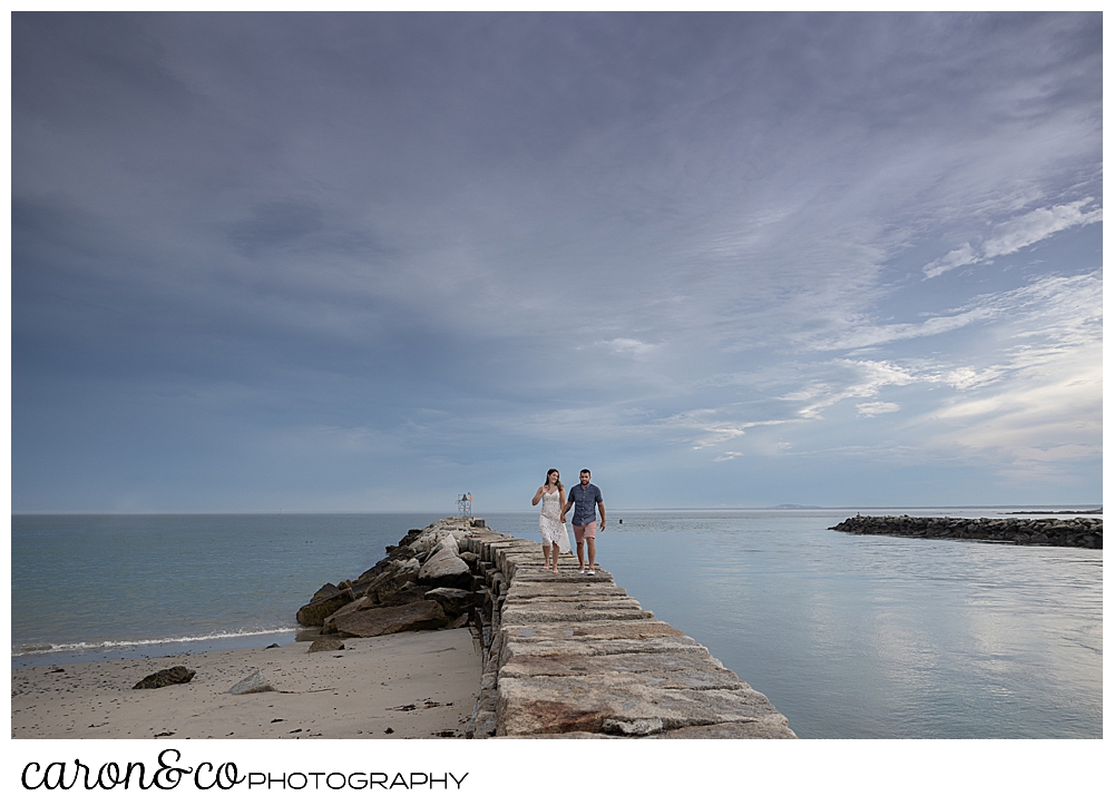 a man and woman walk on the Kennebunkport Maine breakwater, during their Kennebunkport engagement session