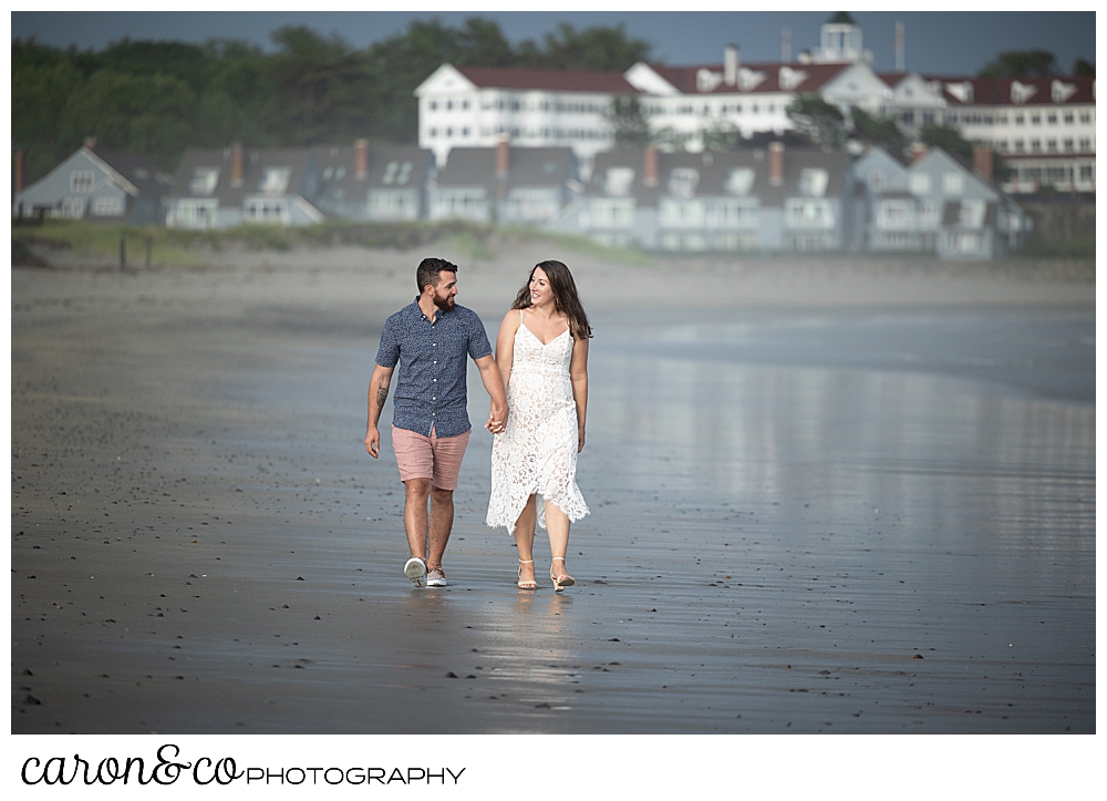 a man and woman hold hands and walk near the water at Gooch's Beach in Kennebunk, Maine, during their Kennebunkport engagement session, the Colony Hotel in the background in Kennebunkport, Maine