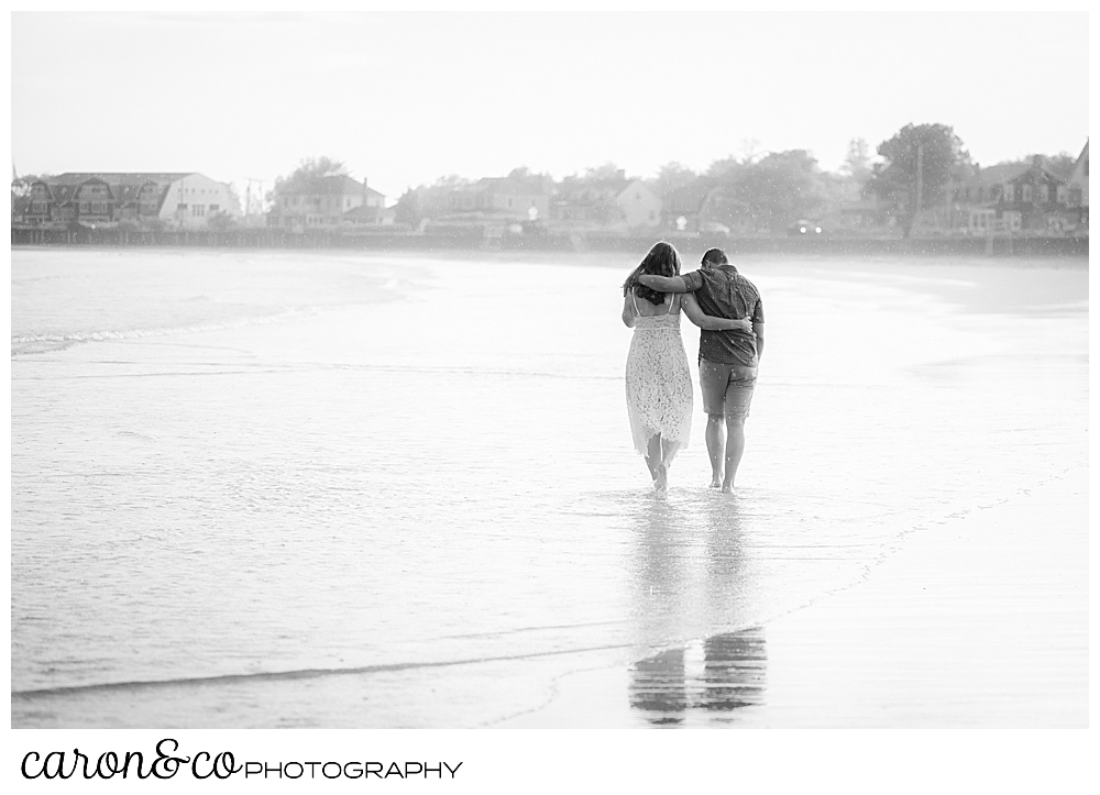 black and white photo of a man and woman, their arms around each other, on Gooch's beach, Kennebunk, Maine, walking away from the camera during their Kennebunkport engagement session