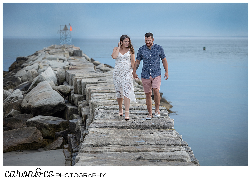 a man and woman walk hand in hand on the Kennebunkport Maine breakwater, during their Kennebunkport engagement session