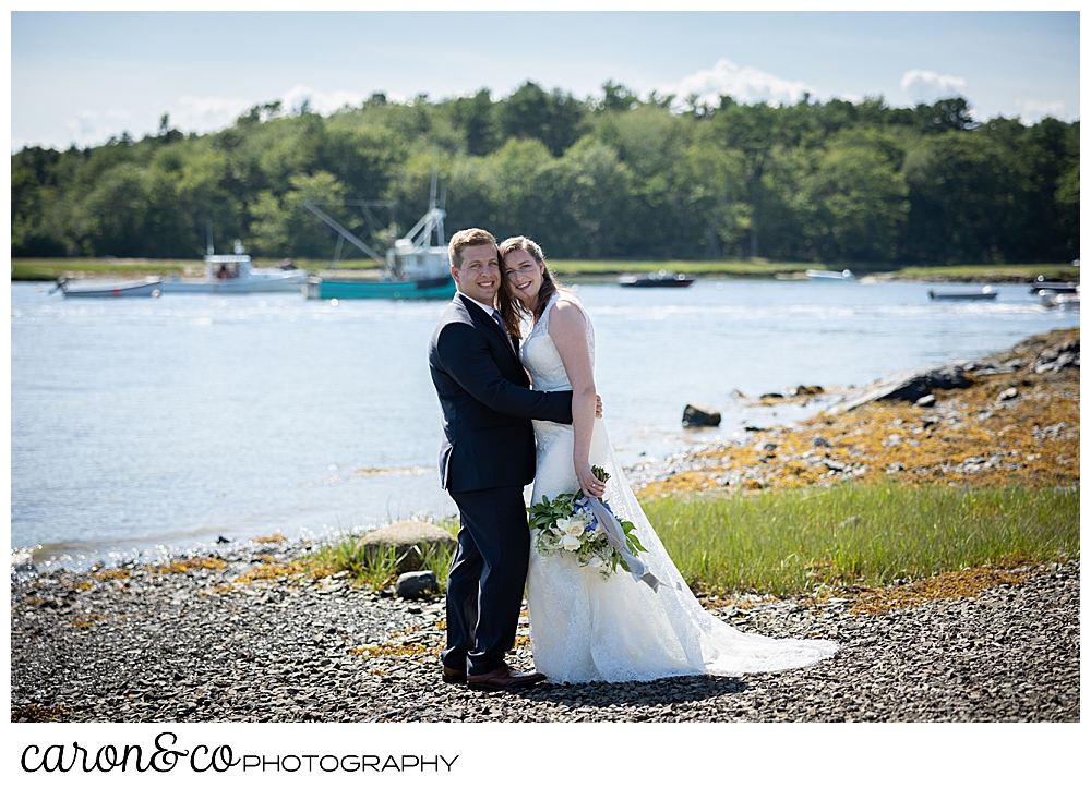 bride and groom hugging each other, on the banks of the Kennebunk River. They're looking at the camera