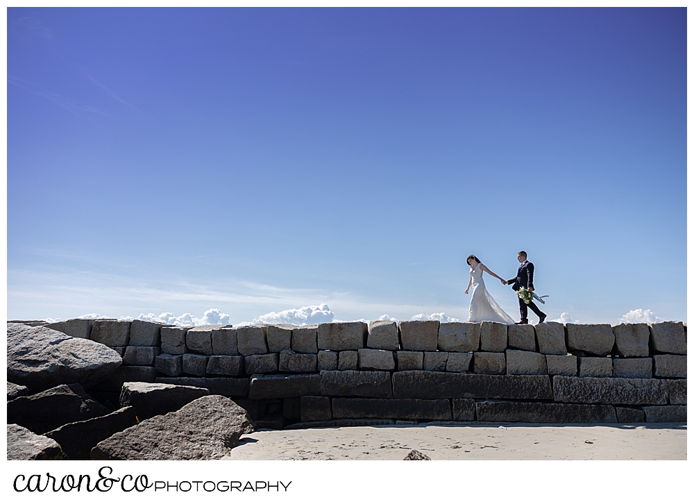 a bride and groom walk along the top of the Kennebunkport Breakwater, holding hands