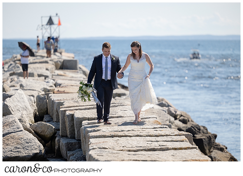 a bride and groom walk on the top of the Kennebunkport Breakwater, they're holding hands