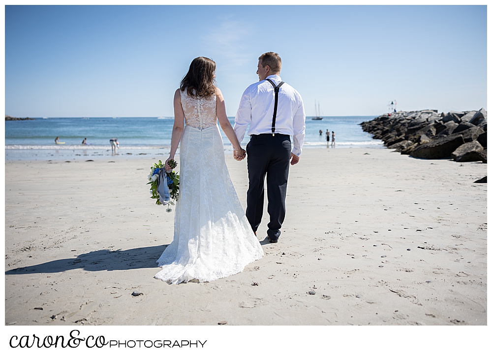 a bride and groom, holding hands, walk away from the camera towards the water at Colony Beach, Kennebunkport, Maine