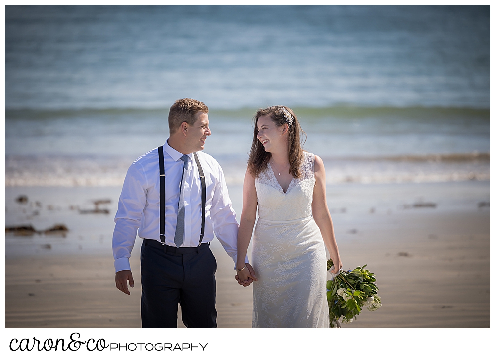 a bride and groom holding hands and walking on the beach, walking towards the camera