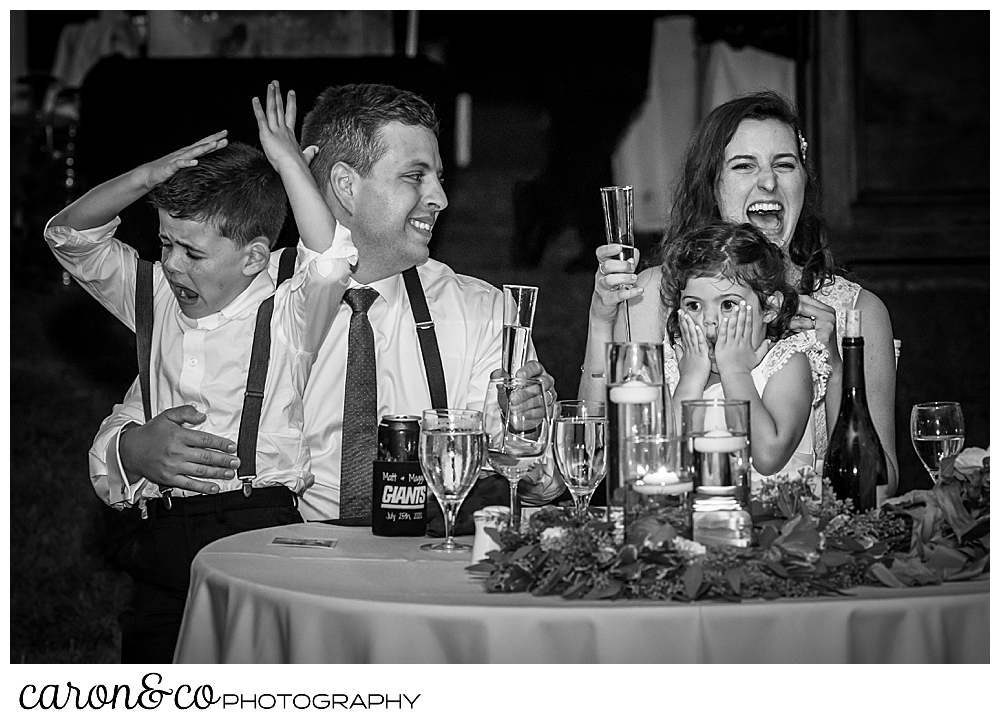 black and white photo of a bride and groom sitting at their sweetheart table, holding a niece and nephew, during toasts at their Nonantum Resort wedding celebration