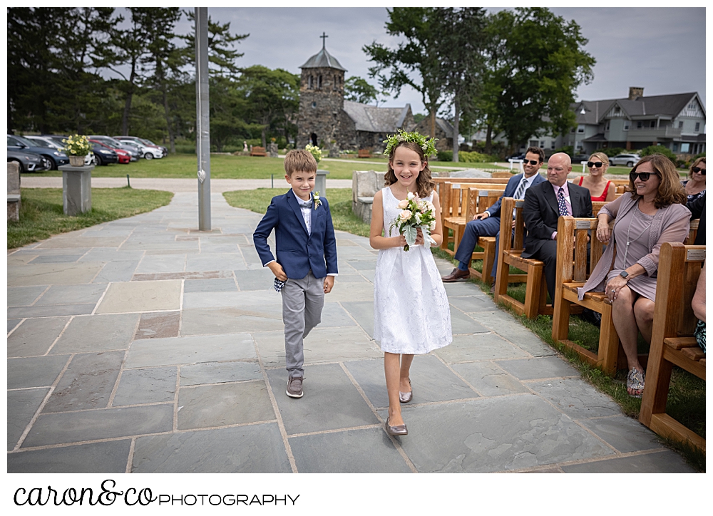 a flower girl and a ring bearer walk down the aisle of the outdoor ceremony site at St. Ann's by-th-sea Episcopal Church, Kennebunkport, Maine