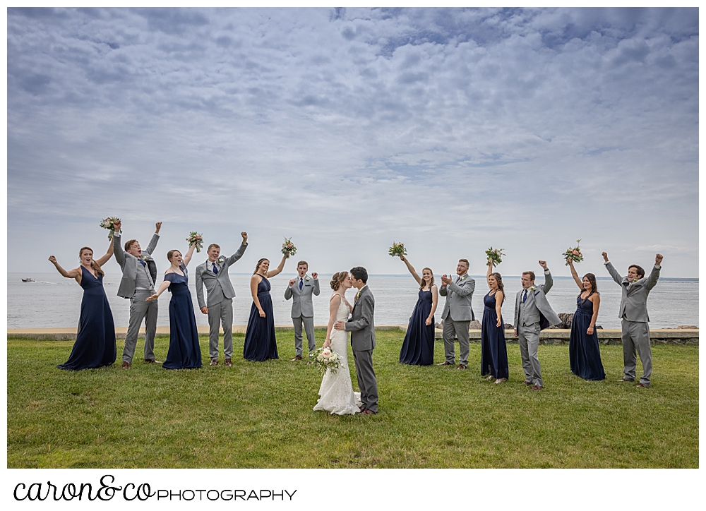 a bride and groom kiss, while their surrounding bridal party cheers, on the lawn at St. Ann's by-the-sea Episcopal Church, Kennebunkport, Maine