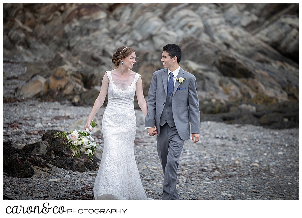 a bride and groom walk hand in hand on a rocky beach in Kennebunkport, Maine