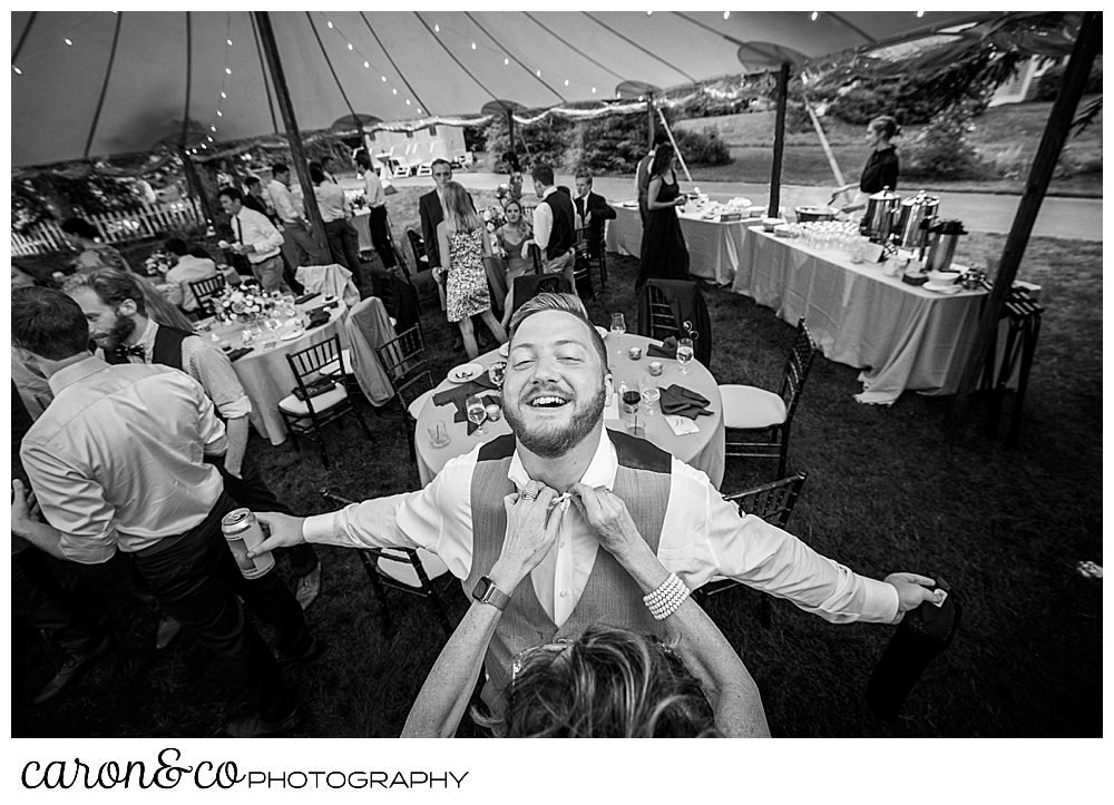 black and white photo of a woman removing a tie from a man who is laughing and holding his arms out during a coastal Kennebunk Maine wedding reception