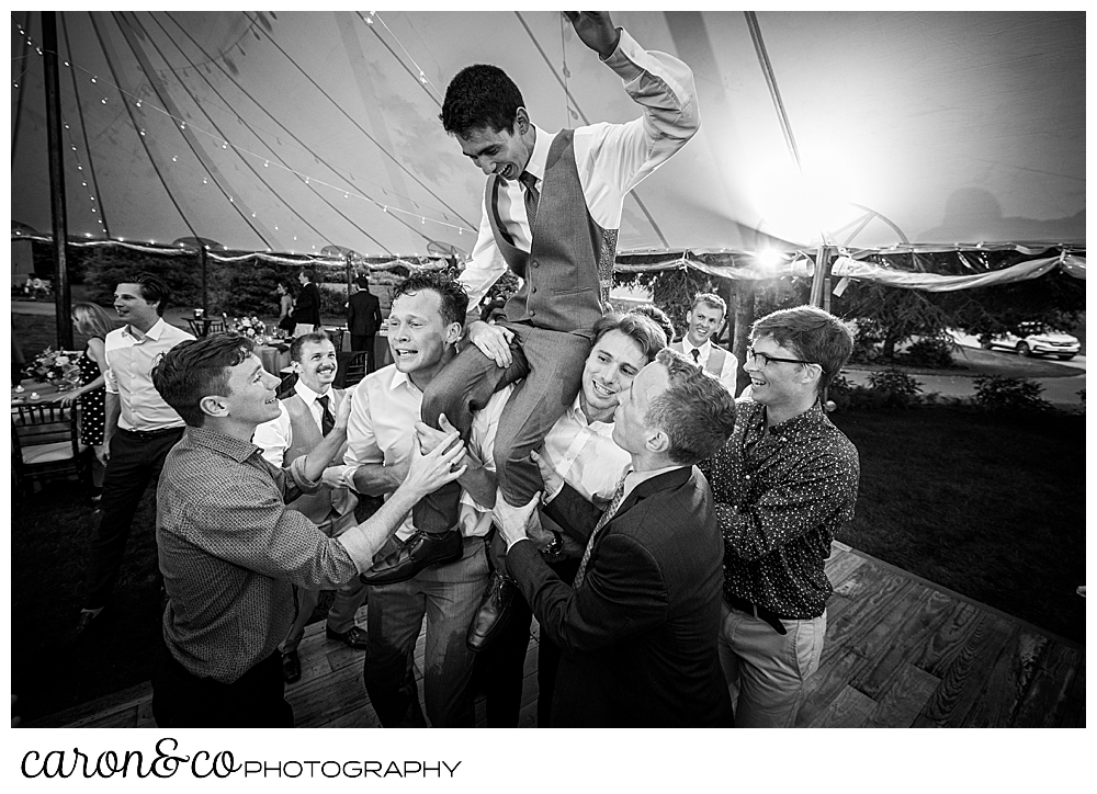 black and white photo of a groom being raised up on his friends shoulders, during a coastal Maine tented wedding