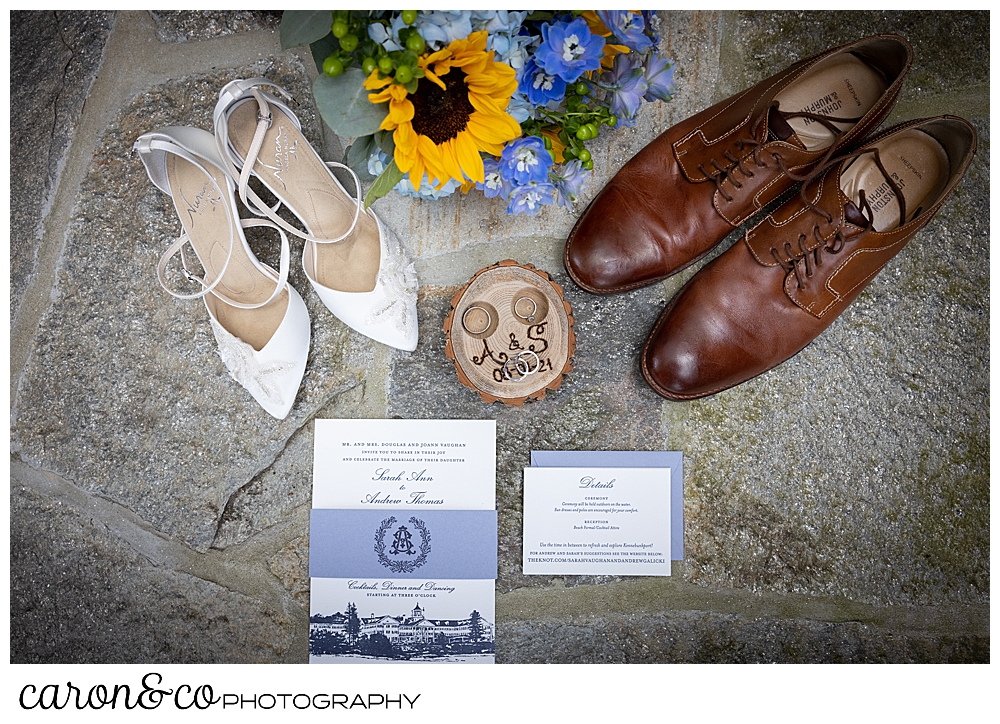 wedding day details: white bridal shoes with straps, mens brown wing tip shoes, a bridal bouquet with blue hydrangea and yellow sun flowers, a blue and white paper suite, and a wooden ring box, all displayed on a slate background