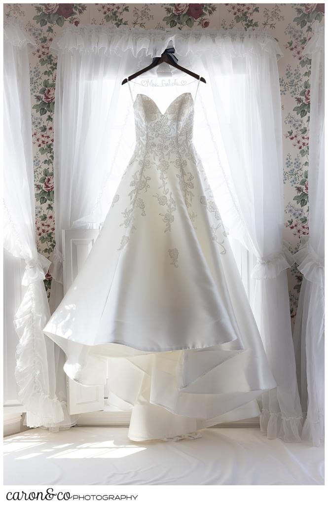 a strapless white wedding dress, with white appliqué flowers, hangs on a wooden hanger in the window of a guest room at the Colony Hotel, Kennebunkport, Maine