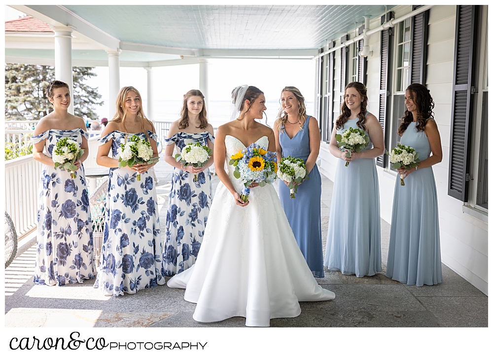 a bride in white, stands in the midst of her bridesmaids, three have light blue dresses, three have blue flowered dresses