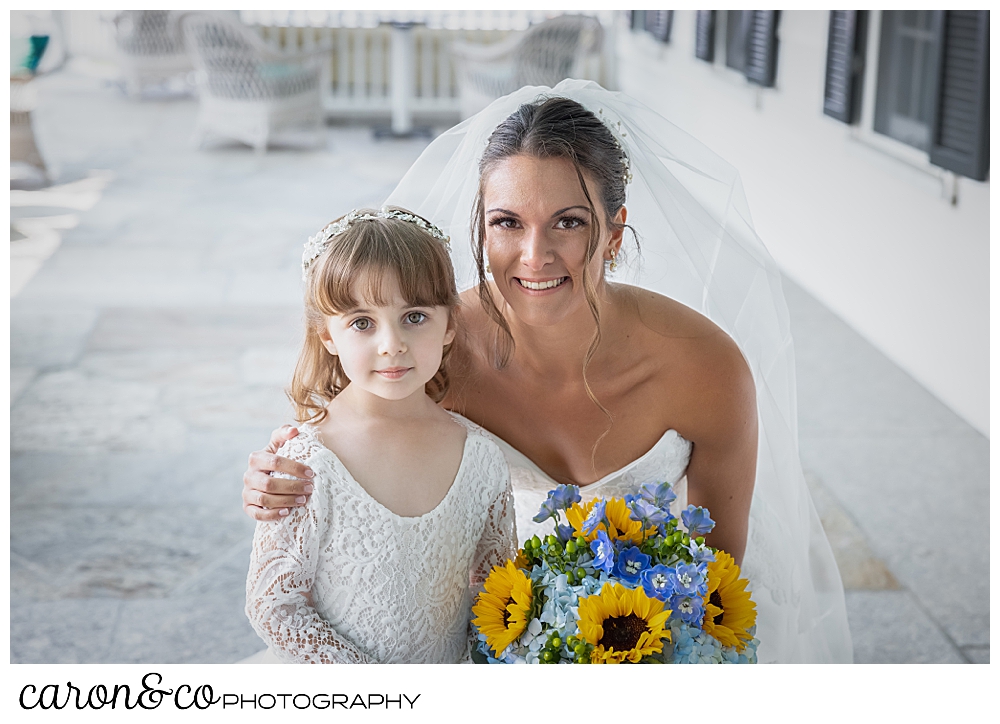 a bride kneels down next to a flower girl, they're both wearing white, the bride has a bouquet of blue hydrangeas and yellow sun flowers