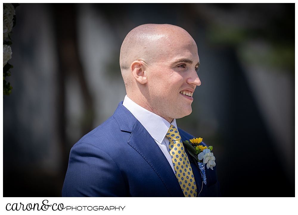 a smiling groom, wearing a blue suit, waits for his bride to come down the aisle at a Kennebunkport wedding at the Colony Hotel