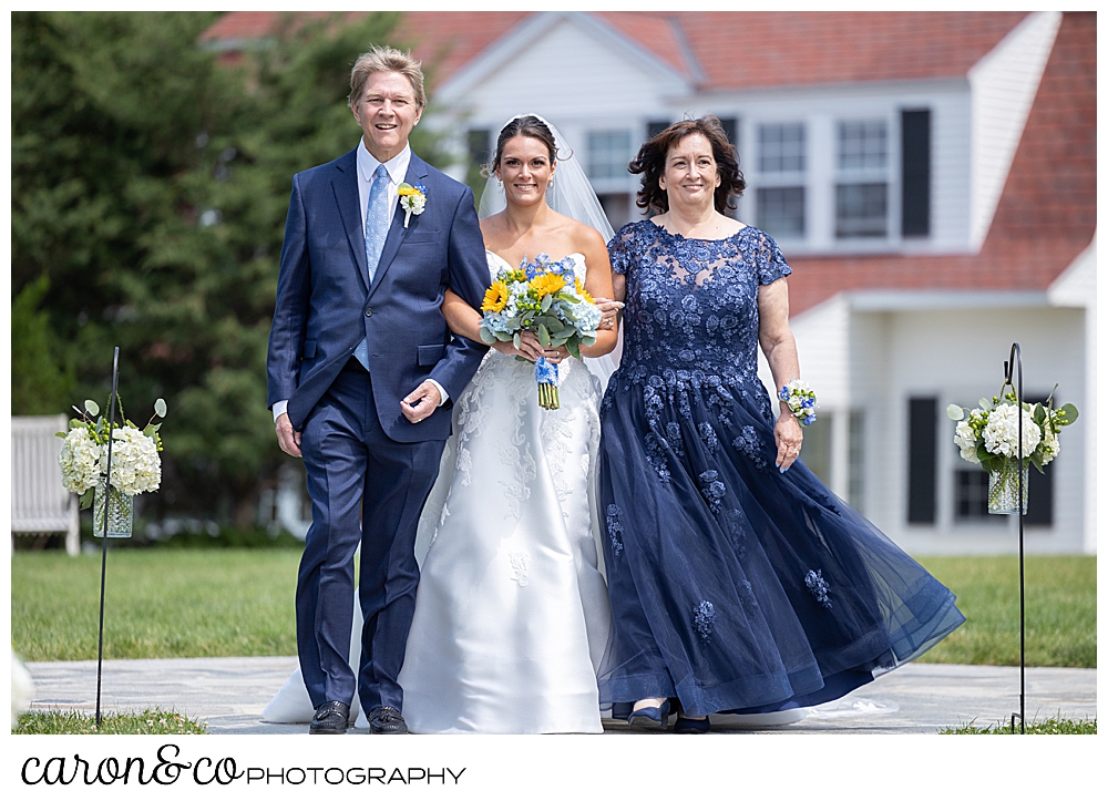 a bride, holding the arms of her mother and father, one on each side, as they walk down the aisle at a Kennebunkport wedding at the Colony Hotel, they're all smiling