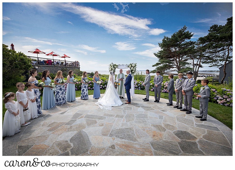 a Colony Hotel outdoor wedding ceremony, with a bride and groom and an officiant in the middle, and 6 bridesmaids and 3 flower girls on the left, and 6 groomsmen on the right