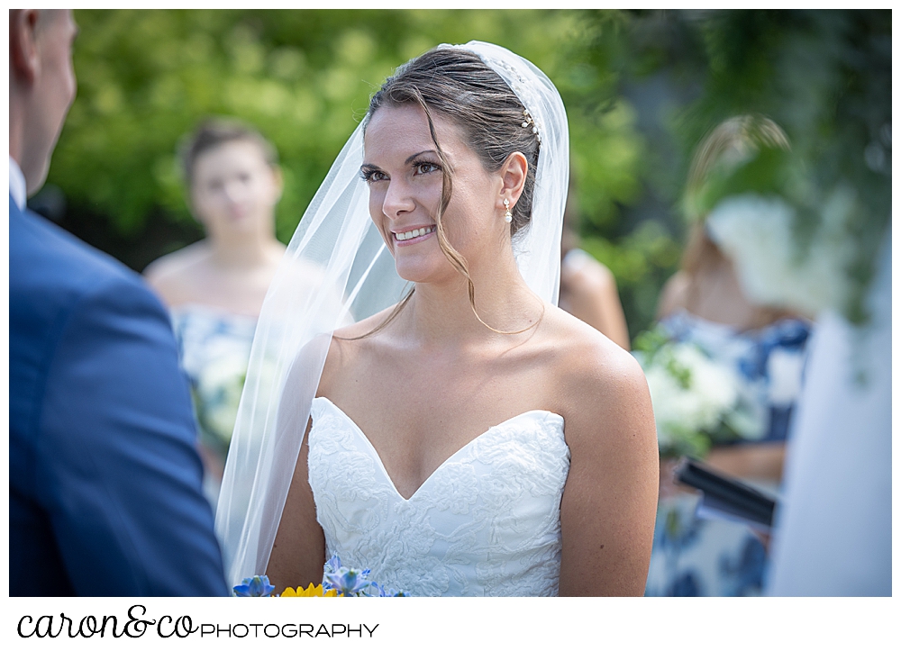 a dark haired, dark eyed bride, wearing a white strapless dress and veil, smiles at her groom during their outdoor ceremony at their Kennebunkport wedding at the Colony Hotel