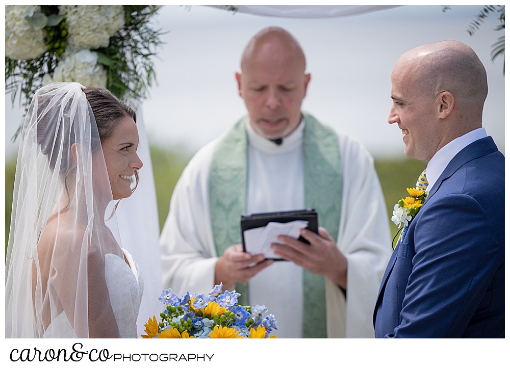 a priest reads while on either side stands a bride and groom, smiling at each other at a Kennebunkport wedding at the Colony Hotel