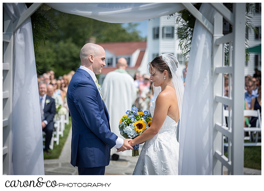 in the fore group are a bride wearing a white dress and veil, and a groom wearing a blue suit, holding hands; in the back ground is the officiant with his back to the camera