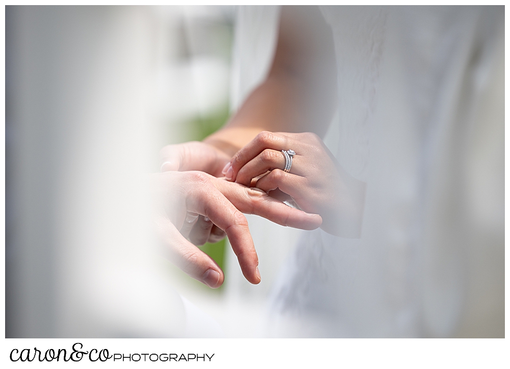 a bride's hand is holding the groom's hand, and she is putting a wedding band on his finger with her other hand