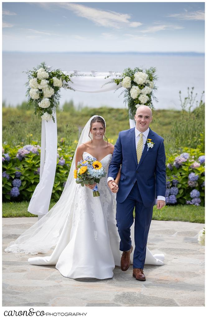 a bride wearing a white dress and veil, carrying a bouquet with blue hydrangeas and yellow sun flowers, is holding the arm of her groom who is wearing a blue suit, during the recessional at their Kennebunkport wedding at the Colony Hotel