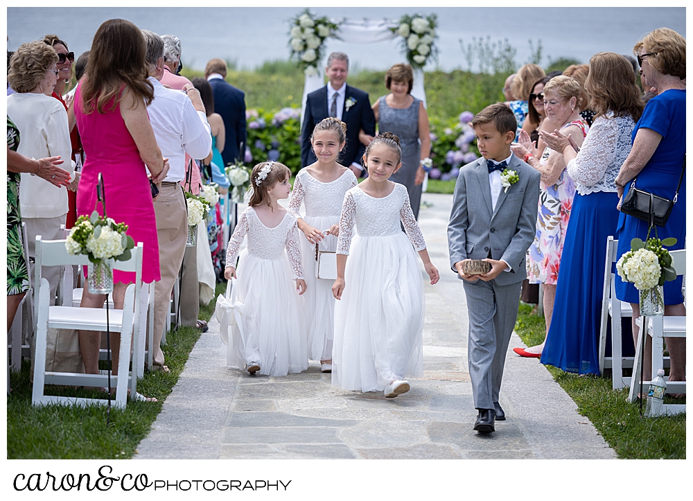 3 flower girls wearing white dresses, and a ring bearer wearing a gray suit are in the fore ground during a Kennebunkport wedding at the Colony Hotel, in the back ground are a women wearing a blue dress and a man wearing a blue suit