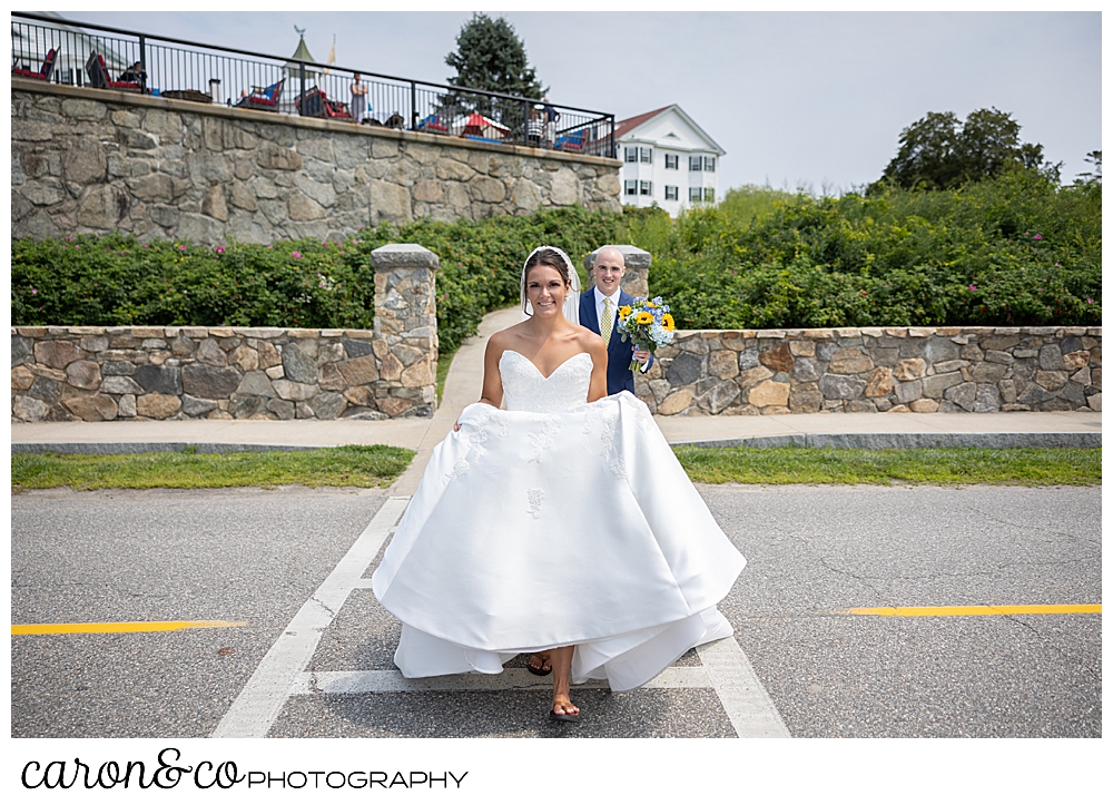 a bride wearing a strapless white dress and a white veil, holds her dress in the front as she crosses the street in front of the Colony Hotel, her groom is following behind, helping with her train, at a Kennebunkport wedding at the Colony Hotel