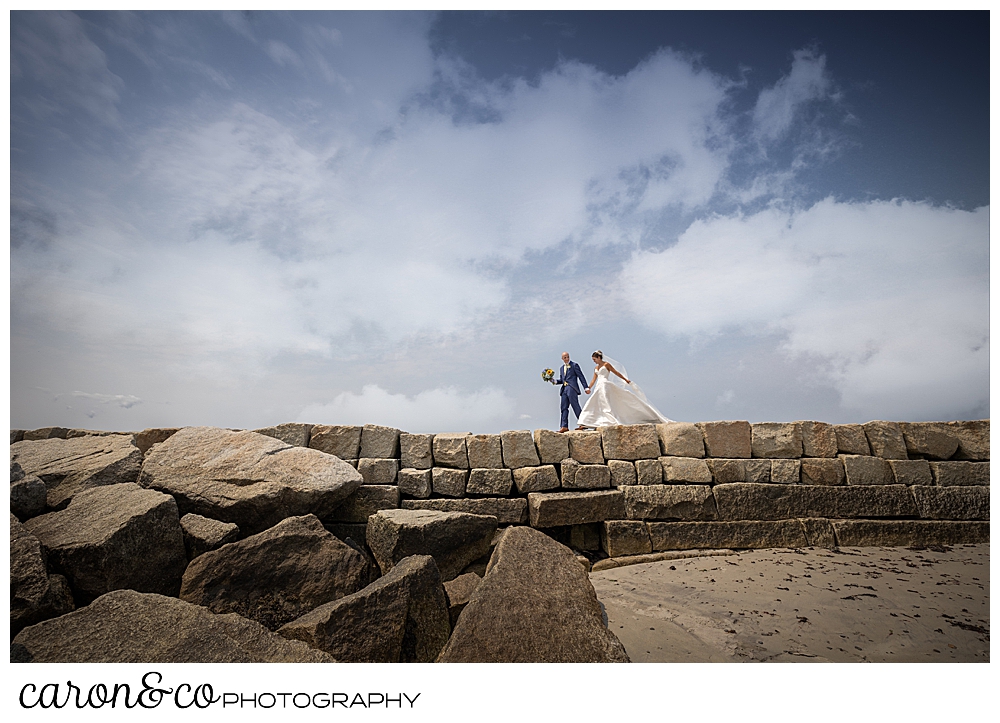 a bride and groom walk along the top of the Kennebunkport Breakwater, the groom is walking ahead of the bride and is holding her bouquet, the clouds and blue sky are in the background