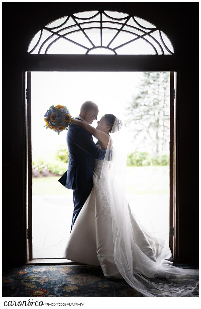 a bride and groom stand in the doorway of the Colony Hotel, their arms around each other, the bride has her bouquet of blue hydrangeas and yellow sun flowers behind the groom's neck