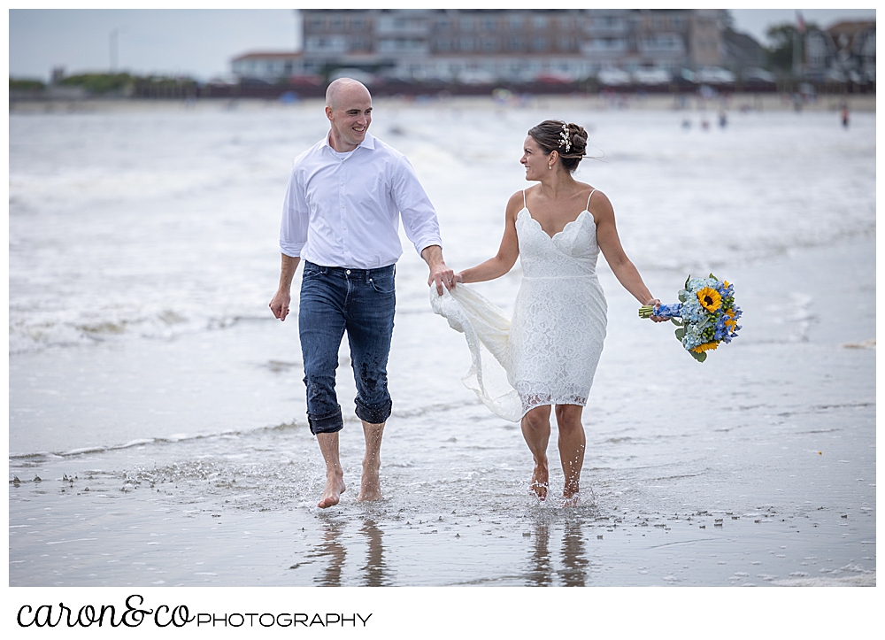 a bride and groom run in the water at Gooch's Beach, Kennebunk, Maine. The bride is wearing a short lace dress and holding a bouquet of blue hydrangeas and yellow sunflowers, the groom is wearing rolled up jeans, and a white shirt