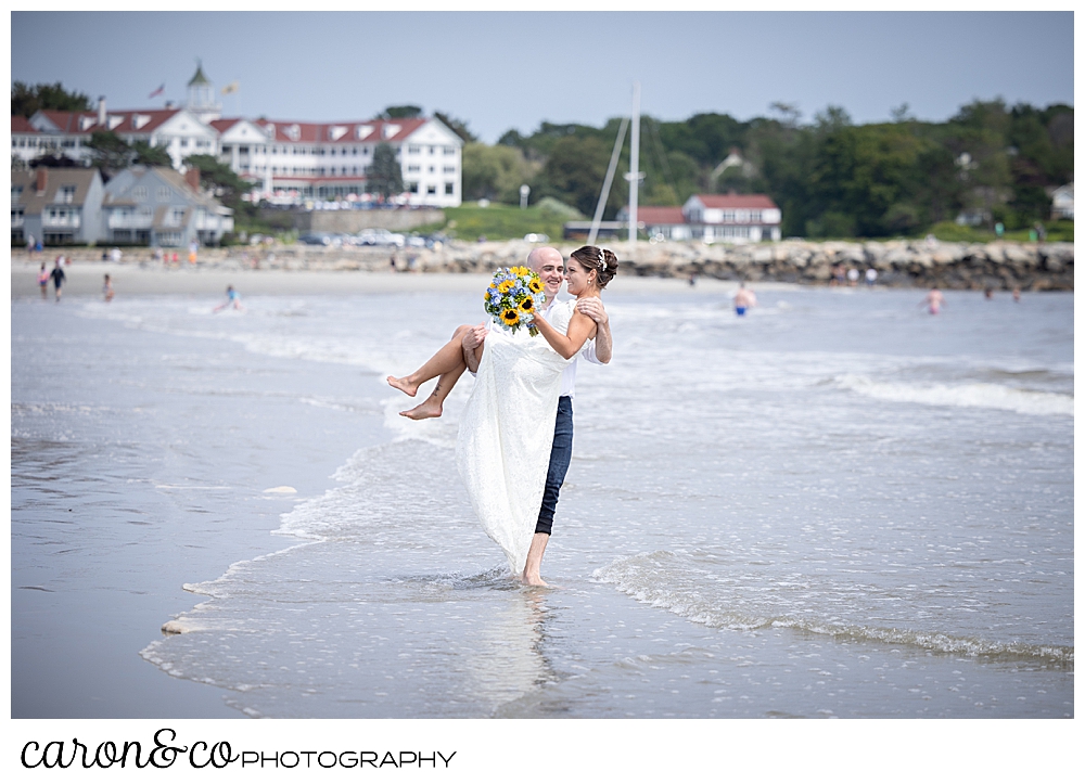 a groom has picked up a bride, and he's walking through the water at Gooch's Beach, the Colony Hotel is in the background