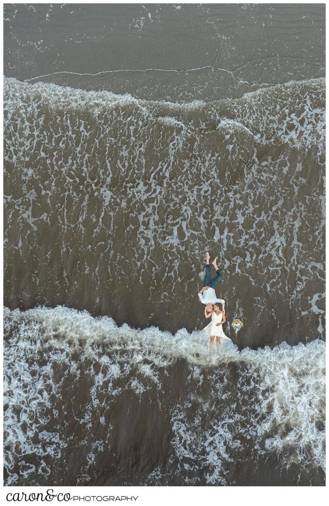 a view of a bride and groom lying down in the water, from a drone. The bride's legs are under the water, as waves come rolling in, Maine drone weddings