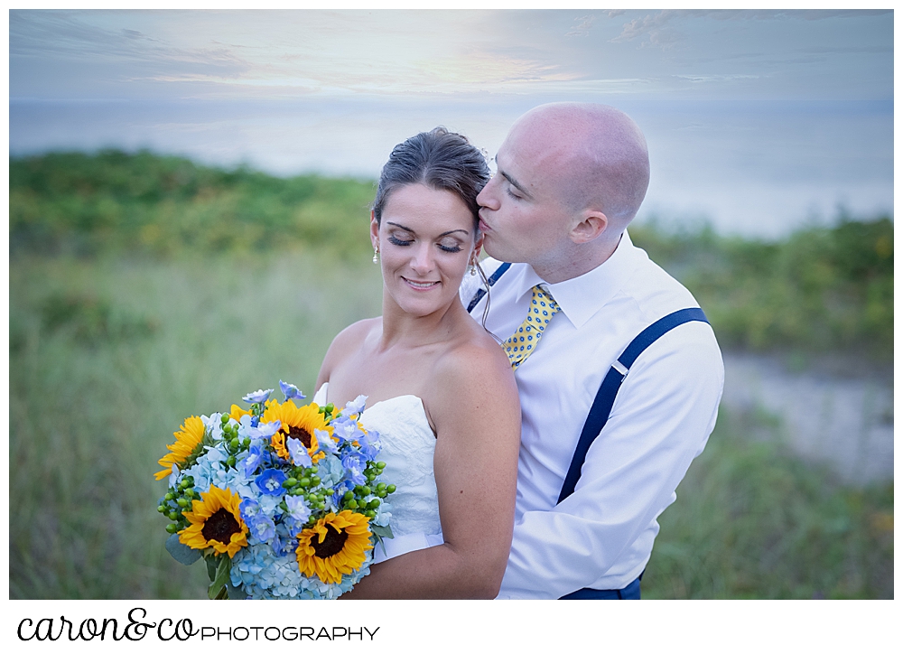 a groom stands behind his bride, kissing her temple, she holds a bouquet of blue hydrangeas and yellow sunflowers, they're in the dunes at Colony Beach, Kennebunkport, Maine