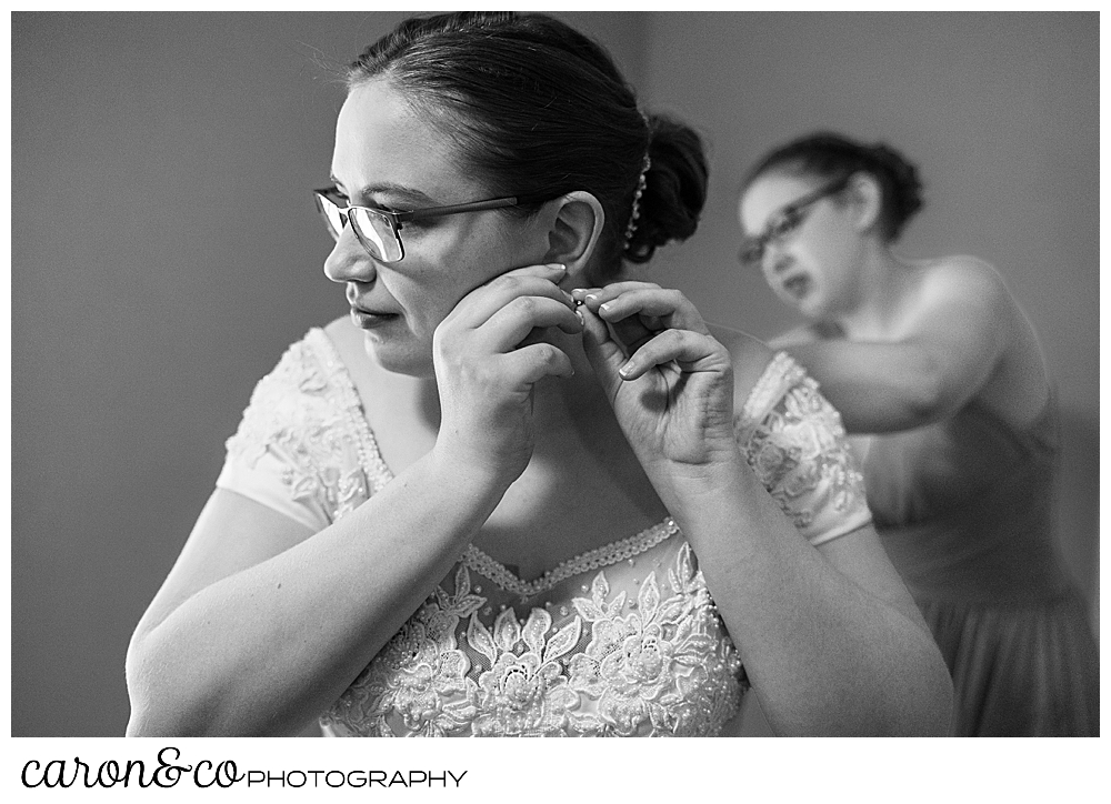 black and white photo of a bride putting her earring on, while her sister helps with her dress in the background