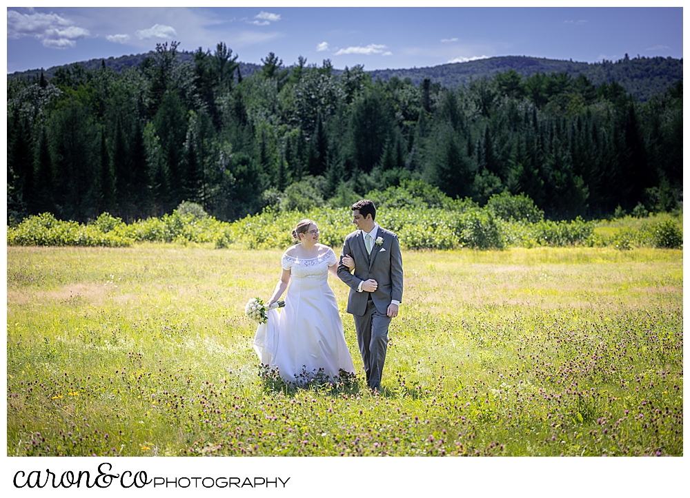 a bride and groom look at each other, while walking in a field, their arms linked